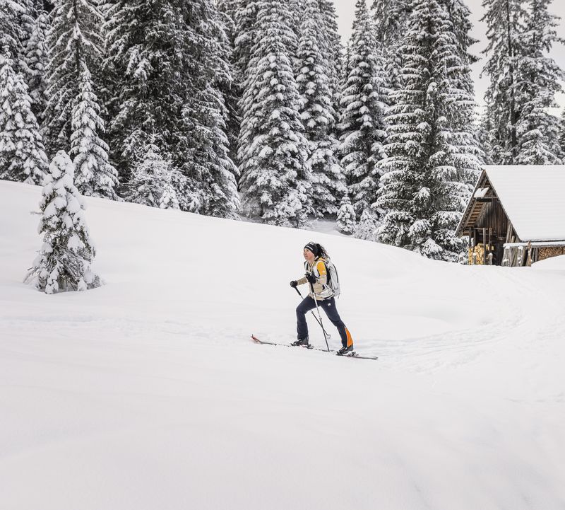 Eine Person beim Skitouren gehen auf einer verschneiten Loipe in der Nähe von Holzhütten, umgeben von schneebedeckten Bäumen.