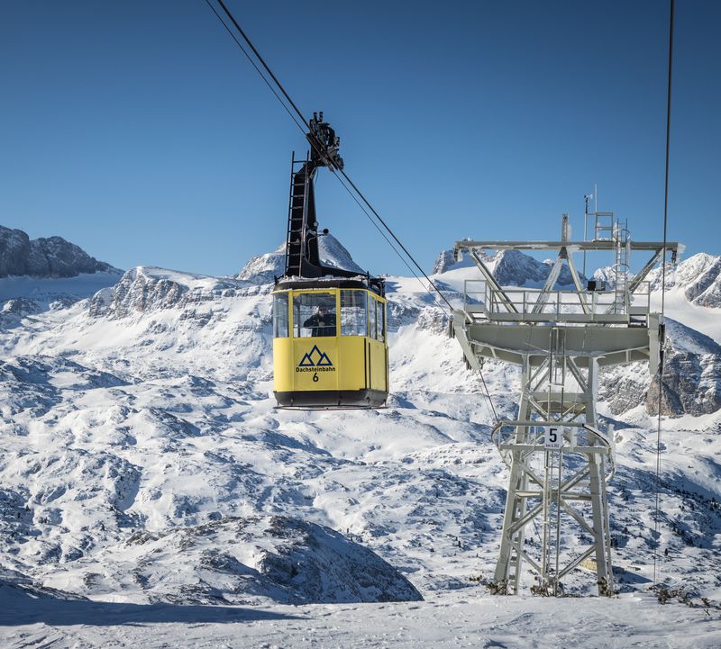 A yellow gondola of the Dachstein Krippenstein cable car floats through the clear winter sky in front of a snow-covered mountain backdrop.
