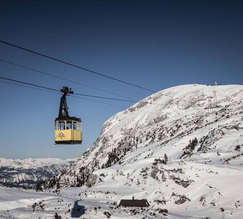 A yellow cable car travels over a snowy mountain landscape under a clear blue sky, with distant peaks visible in the background.