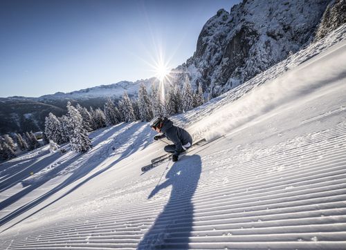 Ein Skifahrer carvt auf einer frisch präparierten Piste, Schnee stiebt auf. Helle Sonne strahlt über verschneiten Bergen und Bäumen.