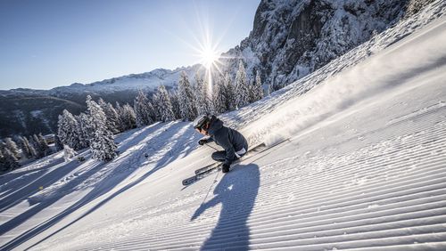Ein Skifahrer carvt auf einer frisch präparierten Piste, Schnee stiebt auf. Helle Sonne strahlt über verschneiten Bergen und Bäumen.