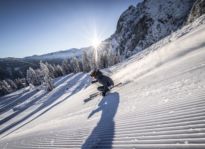 Skifahrer auf frisch präparierter Piste mit aufspritzendem Schnee. Die Sonne strahlt über verschneite Bäume und Berge, lange Schatten.