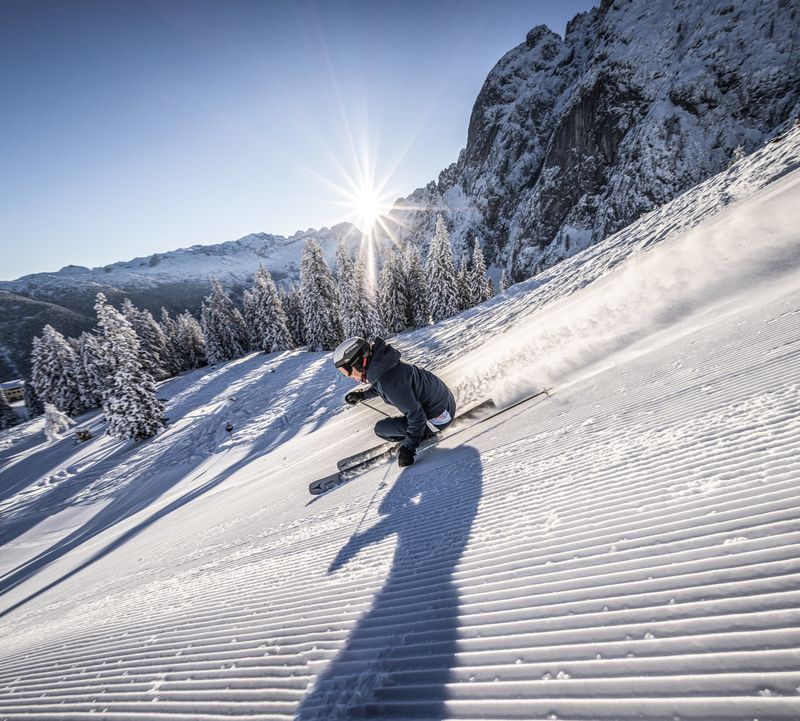 Ein Skifahrer carvt auf einer frisch präparierten Piste, Schnee stiebt auf. Helle Sonne strahlt über verschneiten Bergen und Bäumen.