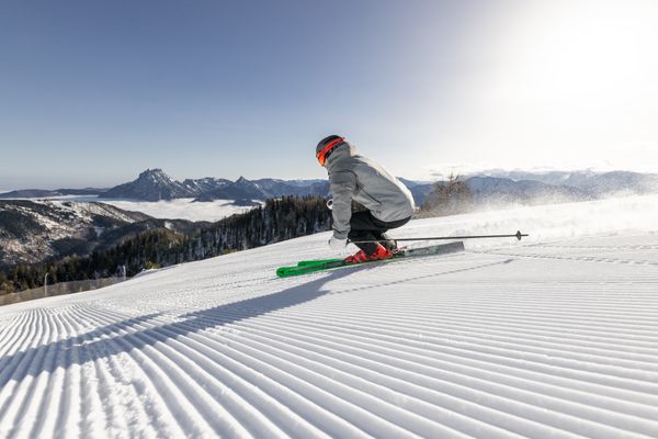 Skifahrer mit grauer Jacke und rotem Helm fährt eine frisch präparierte Piste hinab, im Hintergrund Berge und klarer Himmel – Feuerkogel.