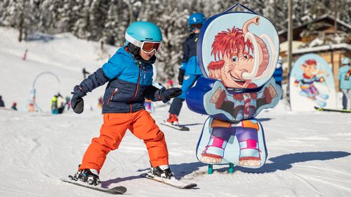 Kind in leuchtend orangefarbener Hose und blauer Jacke beim Skifahren, vorbei an einer Cartoon-Mammut-Figur auf einer verschneiten Piste mit Bäumen im Hintergrund.