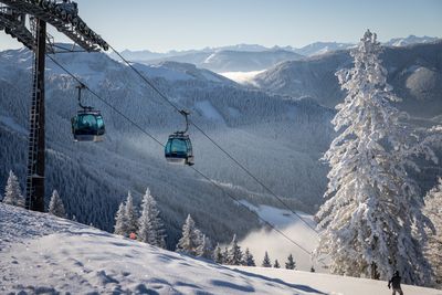 Zwei Gondeln der Donnerkogelbahn schweben über verschneiten Bäumen mit Bergpanorama und blauem Himmel im Hintergrund.