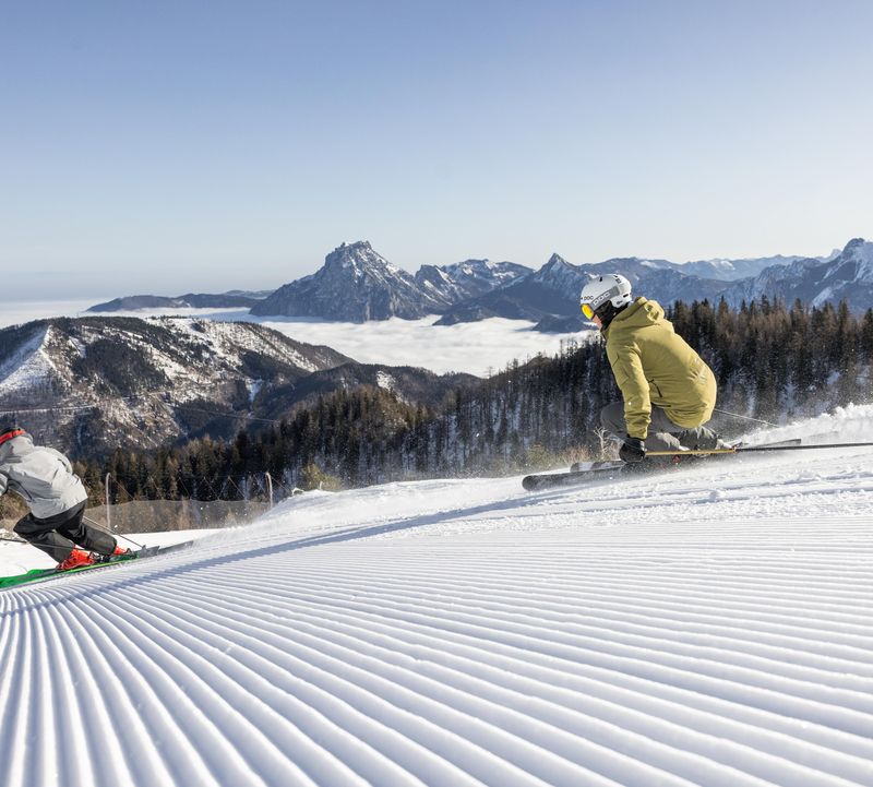 Zwei Skifahrer carven synchron über frisch präparierte Piste mit Blick auf schneebedeckte Berge und Nebelmeer.