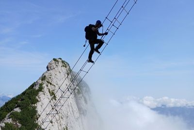 Person climbing a steep, narrow ladder on a mountain with a backdrop of clouds and a clear blue sky.