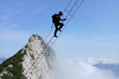 Person, die eine steile, schmale Leiter auf einem Berg hinaufsteigt, mit Wolken und einem klaren blauen Himmel im Hintergrund.