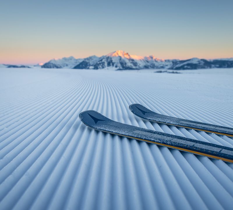 Zwei Skier auf präpariertem Schnee bei Sonnenaufgang, mit fernen Bergen unter klarem Himmel.