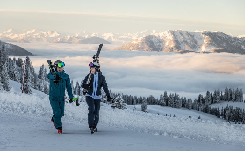 Zwei Skifahrer in Skibekleidung gehen lachend auf der verschneiten Piste mit Skiern auf den Schultern. Im Hintergrund sieht man ein beeindruckendes Bergpanorama mit schneebedeckten Gipfeln und einer Wolkendecke im Tal.