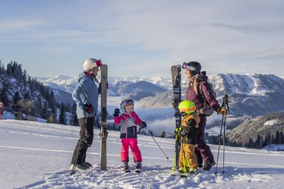 Two women and two children in colorful ski gear are standing on a snow-covered mountainside with skis in hand, with a picturesque mountain range in the background.
