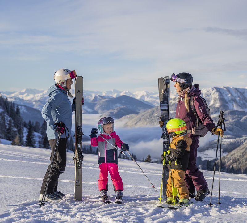 Two women and two children in colourful ski gear stand on a snowy mountainside with skis in hand, with a picturesque mountain range in the background.
