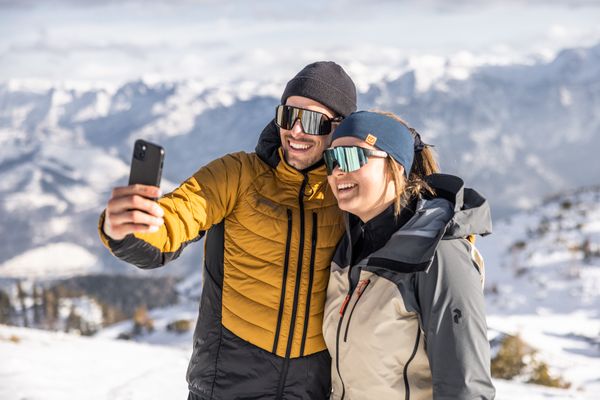 Lächelndes Paar macht Selfie in verschneiter Berglandschaft mit Sonnenbrille und Winterjacke.