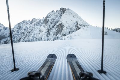 A pair of skis and poles on a freshly groomed snowy slope, with a majestic snow-covered mountain in the background under a clear sky.