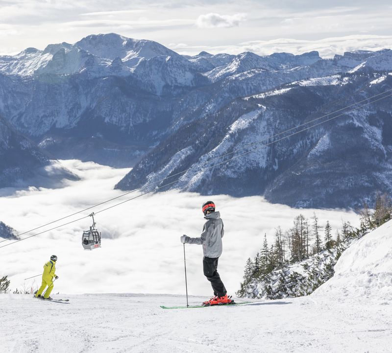 Zwei Skifahrer auf verschneiter Piste vor Bergkulisse, darüber die Gsoll-Bahn über einem wolkenverhangenen Tal – Feuerkogel.