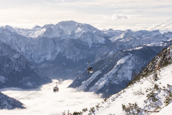 Zwei Gondeln schweben über verschneite Berghänge, unter ihnen liegt ein Tal in dichtem Nebel, umrahmt von steilen Felswänden.