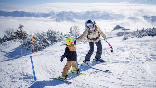 Eine Frau begleitet ein Kind mit grünem Helm beim Skifahren durch einen Slalom auf verschneiter Piste mit Bergblick.