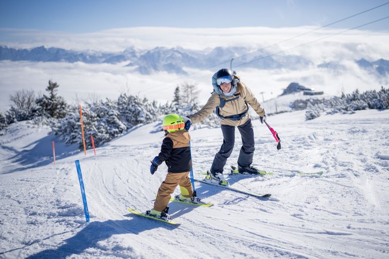 Eine Frau begleitet ein Kind mit grünem Helm beim Skifahren durch einen Slalom auf verschneiter Piste mit Bergblick.