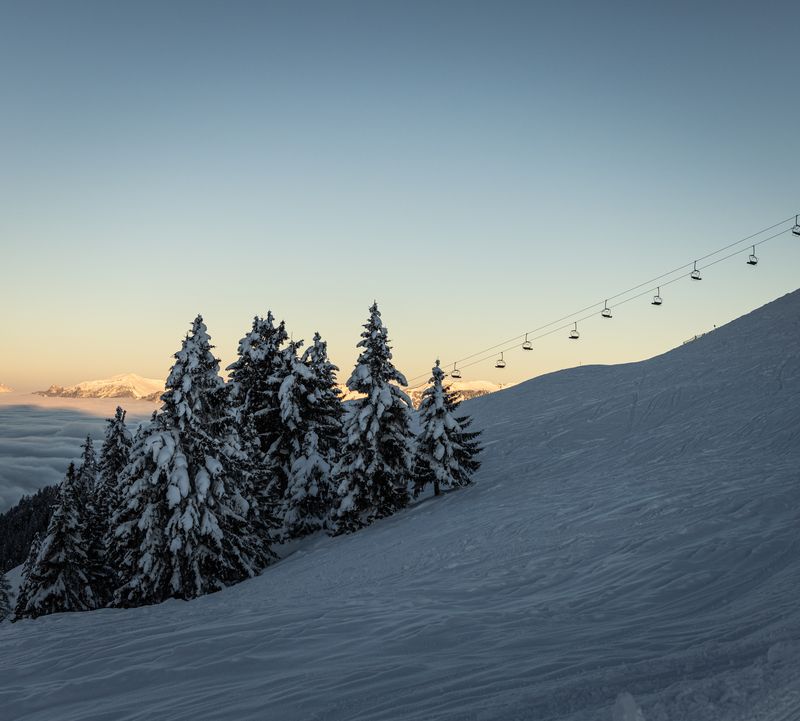 Snow-covered piste with chairlift, snow-covered trees and distant views of the mountains at sunrise.