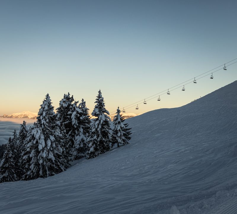 Beschneite Piste mit Sessellift, verschneiten Bäume und Fernblick auf die Berge bei Sonnenaufgang.er a clear sky at sunset.