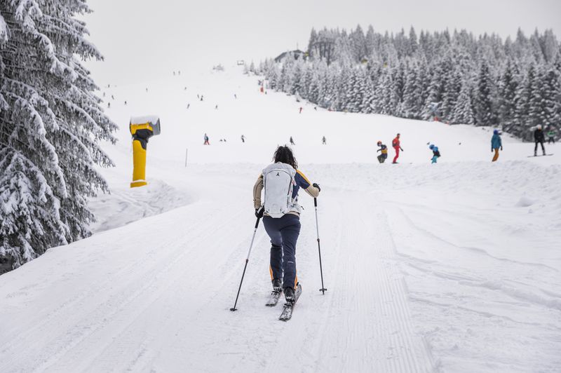 Eine Person geht eine verschneiten Piste bergauf, umgeben von schneebedeckten Bäumen und anderen Skifahrern in der Ferne unter einem bedeckten Himmel.