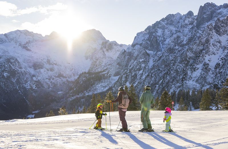 A family of four, dressed in colorful ski gear, stands on a snowy slope with majestic mountains in the background under a bright sun.