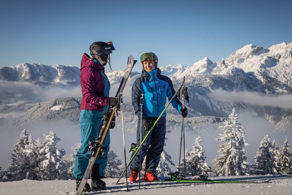 Two skiers in winter gear stand with skis on a snowy mountain, surrounded by snow-covered trees and mountains under a clear blue sky.