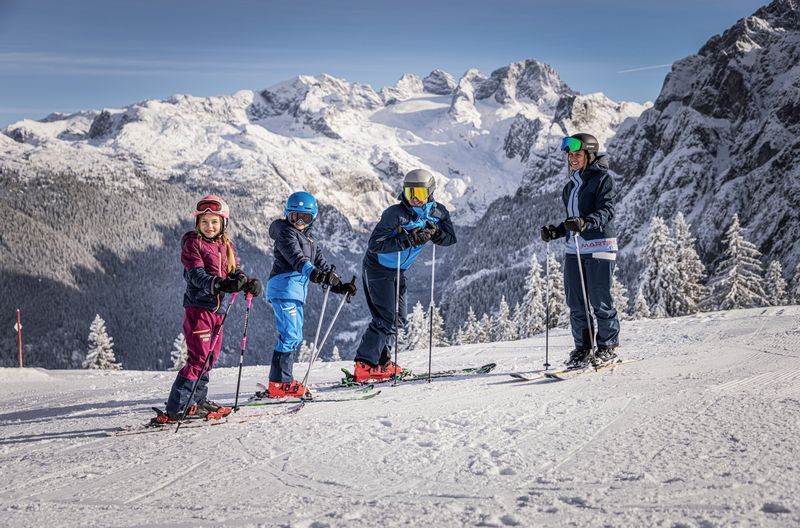 4 Köpfige Familie in Skiausrüstung stehen auf einer verschneiten Piste mit Dachstein im Hintergrund.
