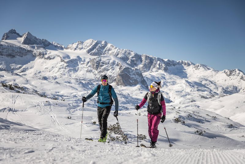 Two people hike on touring skis through the snowy Dachstein mountains under a clear blue sky