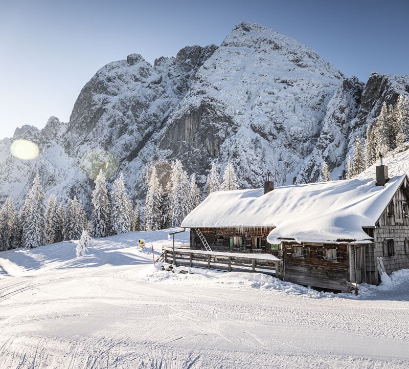 Snow-covered hut in a mountain landscape, surrounded by snow-covered trees under a clear blue sky illuminated by the sun.