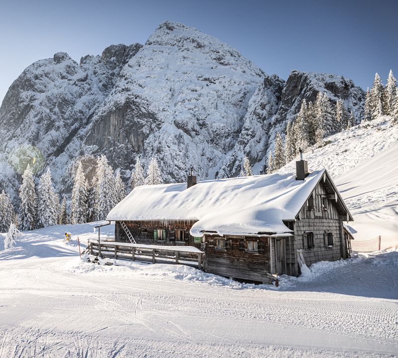 Verschneite Hütte in einer Berglandschaft, umgeben von schneebedeckten Bäumen unter einem klaren blauen Himmel, der von der Sonne erhellt wird.