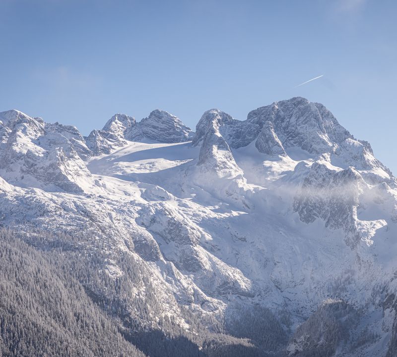 Blick auf den schneebedeckten Dachstein Geltscher mit markanten Gipfeln, umgeben von einem verschneiten Wald unter einem strahlend blauen Himmel.