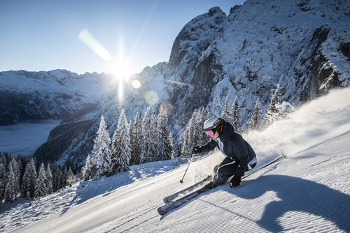 Eine Skifahrerin in dunkelblauer Ausrüstung und weißem Helm fährt einen verschneiten Berghang hinunter, umgeben von schneebedeckten Bäumen, während die Sonne hell hinter den Gipfeln hervor kommt.