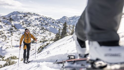 Ein Skitourengeher steigt lächelnd durch das verschneite Gelände, im Vordergrund sieht man Skischuhe und Ski einer zweiten Person.