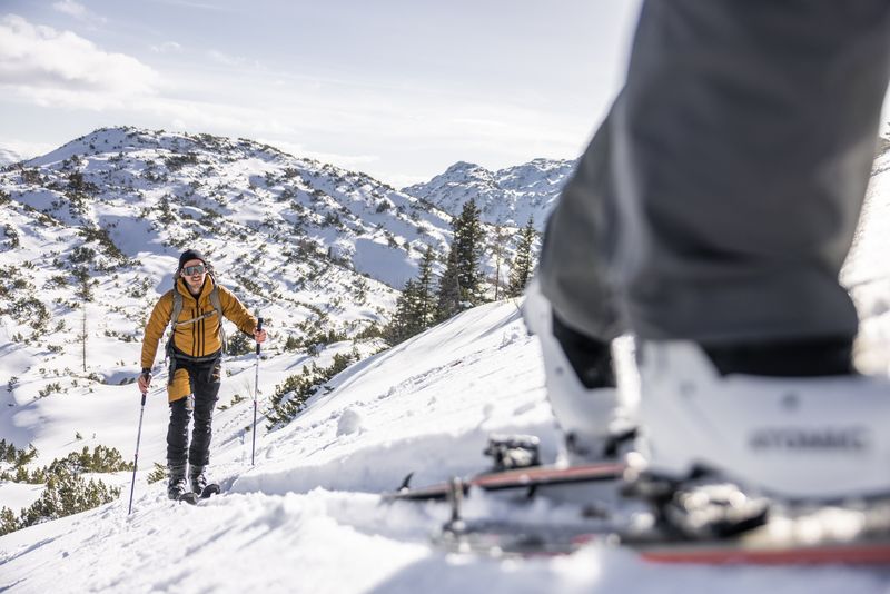 Ein Skitourengeher steigt lächelnd durch das verschneite Gelände, im Vordergrund sieht man Skischuhe und Ski einer zweiten Person.