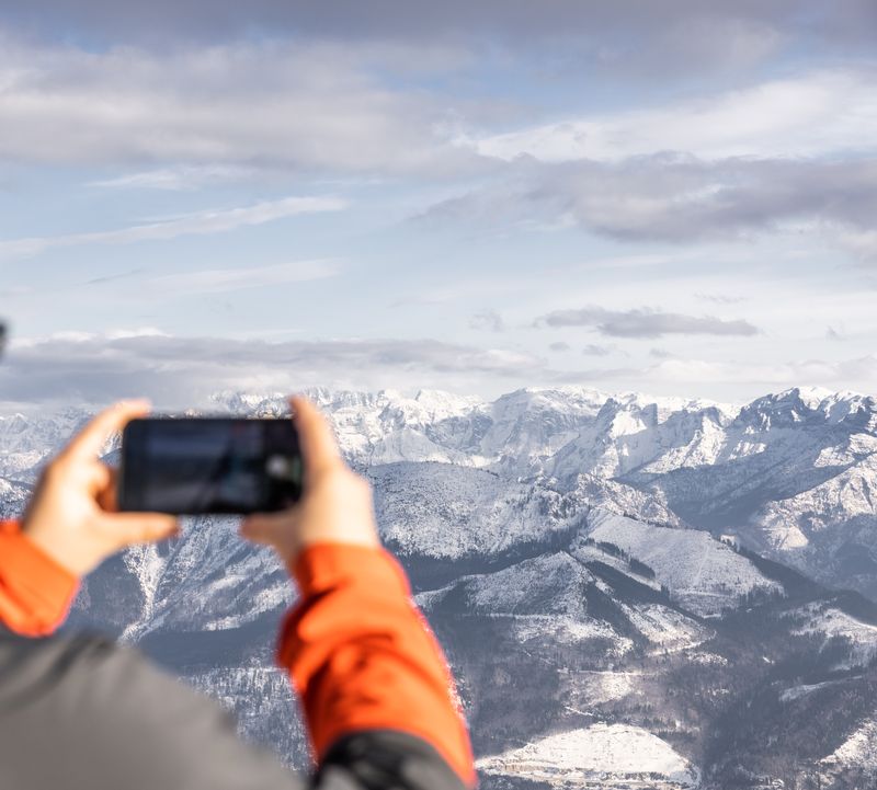 Woman wearing a headband and ski jacket holds her smartphone up to a snow-covered mountain range with peaks.