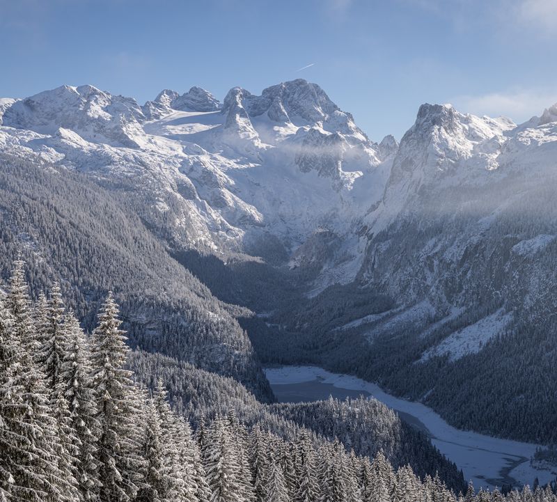 Winterlicher Anblick auf das schneebedeckte Dachstein Massiv mit umliegenden Gipfeln, verschneiten Wäldern und einem zugefrorenen See im Tal.