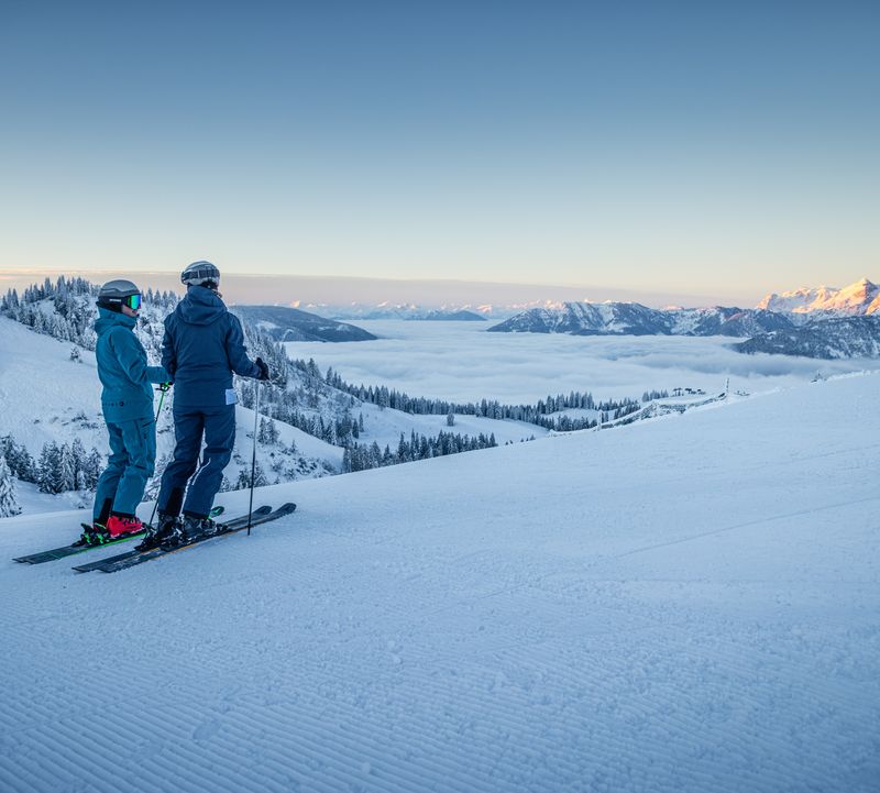 Zwei Skifahrer auf einer Piste blicken auf ein Wolkenmeer im Tal und sonnenbeschienene Bergspitzen.