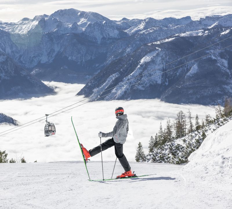Skier in grey jacket and red ski boots stands with skis on a snowy slope, mountains and clouds behind him - Feuerkogel.