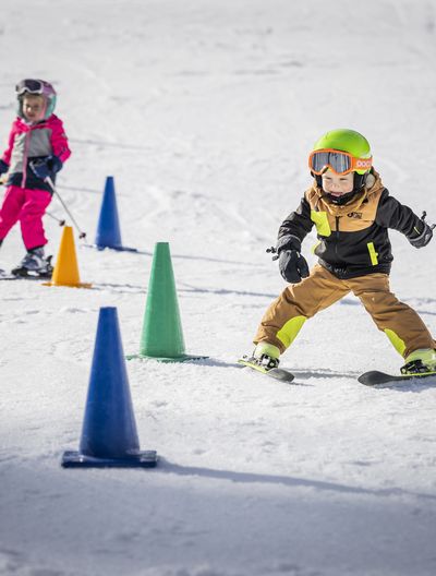 Zwei Kinder in bunter Skikleidung fahren mit Helm und Skibrille auf Schnee und manövrieren um bunte Kegel, die in einem Slalom aufgebaut sind.