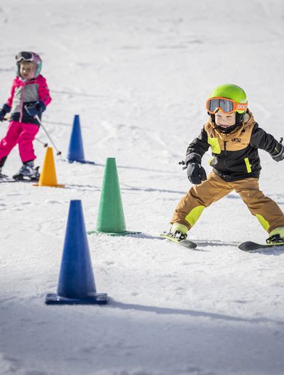 Two children in colourful ski clothing with helmets and ski goggles ski on snow and manoeuvre around colourful cones set up in a slalom.