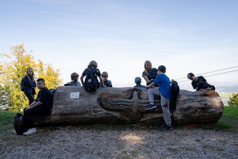 Kinder und eine Frau sitzen und stehen auf einem großen Baumstamm mit Adlerschnitzerei im Freien unter blauem Himmel.