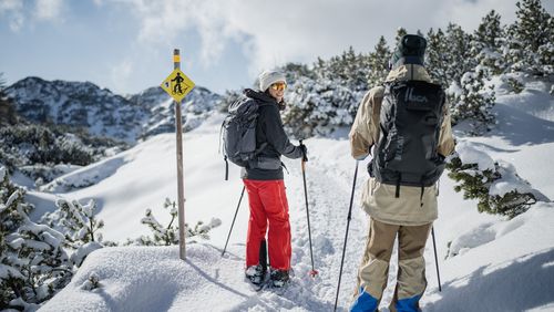 Zwei Personen auf einem verschneiten Winterwanderweg, eine lächelt zurück, umgeben von Bergen und Latschen.