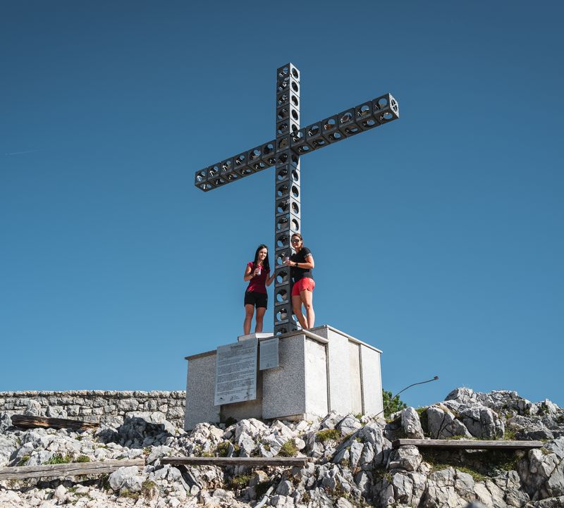 Zwei Frauen in Sportkleidung posieren beim Europakreuz am Gipfel des Feuerkogels unter blauem Himmel.