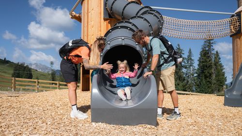 Eine Familie spielt auf einem Abenteuerspielplatz in den Bergen. Ein kleines Mädchen rutscht lachend aus einer Röhrenrutsche, während zwei Erwachsene zusehen.
