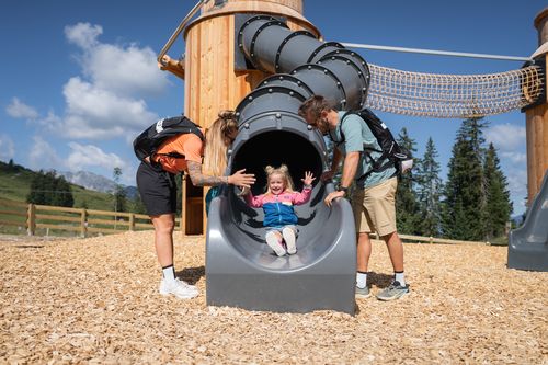 Eine Familie spielt auf einem Abenteuerspielplatz in den Bergen. Ein kleines Mädchen rutscht lachend aus einer Röhrenrutsche, während zwei Erwachsene zusehen.