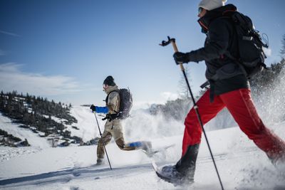 Zwei Schneeschuhwanderer stapfen dynamisch durch tiefen Schnee, Schnee wirbelt auf, klarer Himmel im Hintergrund.