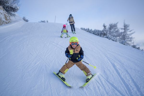 Ein Kind in heller Skikleidung fährt unter strahlend blauem Himmel einen verschneiten Hang hinunter, gefolgt von einem weiteren Kind und einem Erwachsenen.