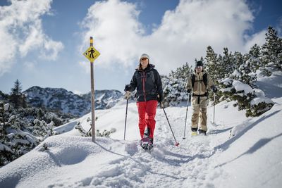 Zwei Personen wandern mit Schneeschuhen an einem gelben Routenschild vorbei durch sonnige Schneelandschaft mit Bergkulisse.
